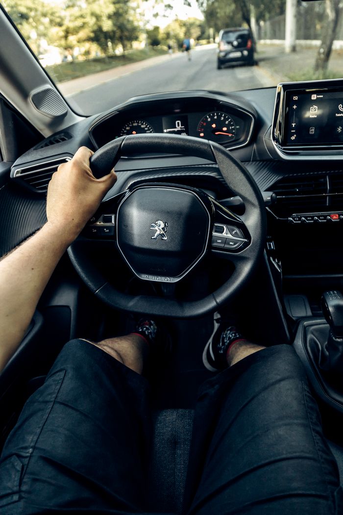 gallery-3 View of a person driving a Peugeot car, focused on the steering wheel and dashboard in a lush green setting.