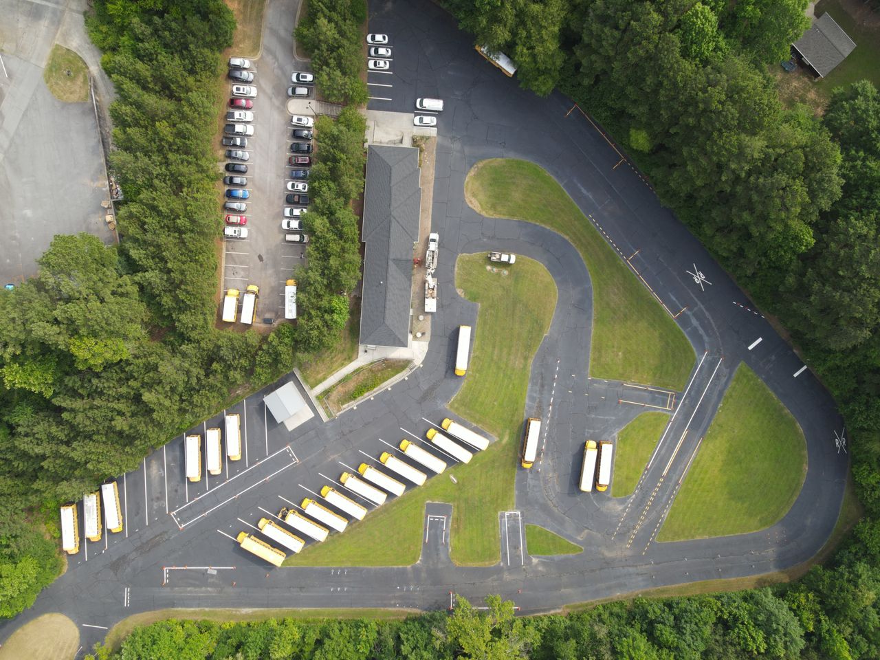 about-01 Aerial shot of a school parking lot with multiple yellow school buses and parked cars surrounded by trees.